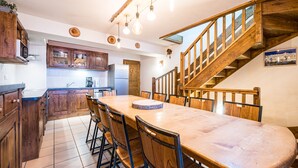 Een keuken met houten kasten, een eettafel met stoelen, een koelkast en een houten trap.