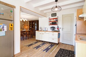 A modern kitchen with white cabinets, a built-in oven, a refrigerator, a stone wall, a wooden dining table, and a hanging light fixture. - La Maison de Gianni - 7km de la Plage (Quettreville-sur-Sienne)