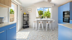 A kitchen with blue cabinets, a white countertop, a dining table with chairs, a black shelf, and a window with a view of trees. - Kerblue - Charmante maison à 3 km de la plage (Carnac)