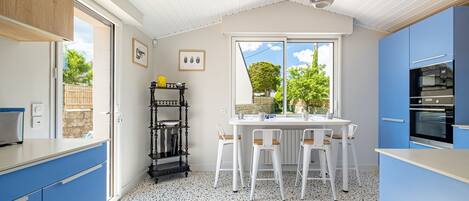 A kitchen with blue cabinets, a white countertop, a dining table with chairs, a black shelf, and a window with a view of trees.