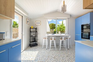 Ett kök med blå skåp, en vit bänkskiva, ett matbord med stolar, en svart hylla och ett fönster med utsikt över träd.