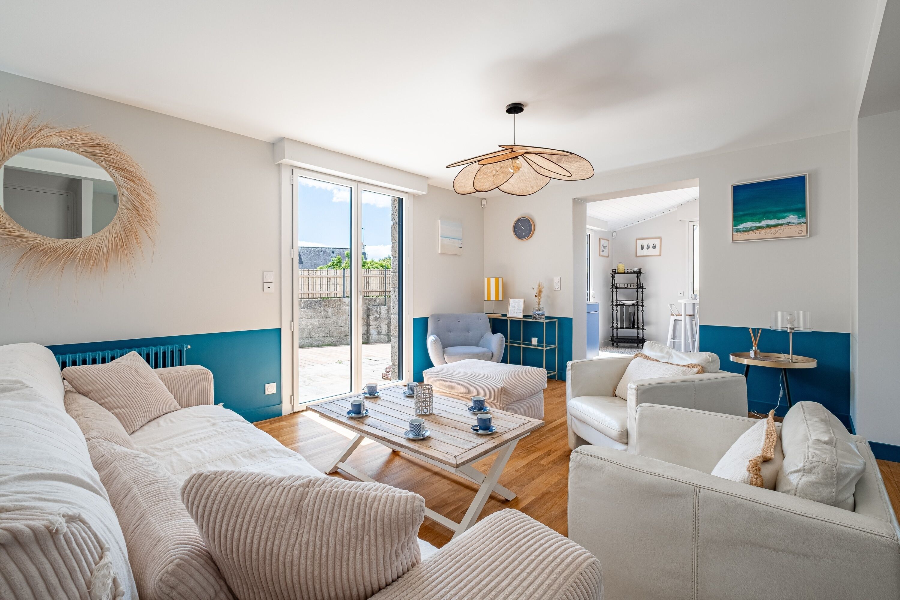 Un salon moderne comprenant un canapé, des fauteuils, une table basse et une table à manger. On y trouve également un grand miroir, un luminaire suspendu et une vue sur l’extérieur grâce à une porte coulissante.