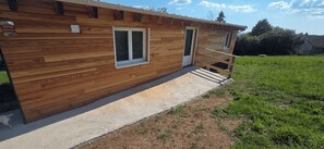 Une cabane en bois avec deux fenĂȘtres, une porte et un chemin en bĂ©ton menant Ă une terrasse en bois.