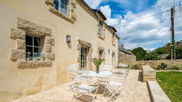 Eine Terrasse mit einem runden Tisch und Stühlen, eine Steinmauer und ein Gebäude mit Fensterrahmen aus Stein.