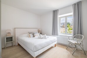 A bedroom with a bed, bedside tables, a chair, a window with curtains, and a view of the outside. - Longère Kerlescan - À 5 km de la plage (Carnac)