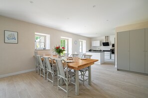 A spacious dining area with a wooden table and chairs, a kitchen in the background, and a large window providing natural light. - Longère Kerlescan - À 5 km de la plage (Carnac)