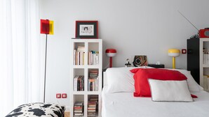 A neatly arranged bedroom with a white bed, a bookshelf, a red and white lamp, and a red and yellow floor lamp. - Atelier des Harmonies - L'atelier d'artiste au cœur de Lyon (Lyon)