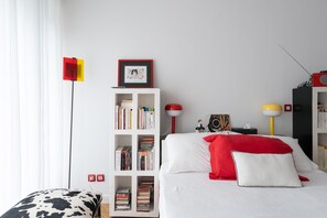 A neatly arranged bedroom with a white bed, a bookshelf, a red and white lamp, and a red and yellow floor lamp. - Atelier des Harmonies - L'atelier d'artiste au cœur de Lyon (Lyon)