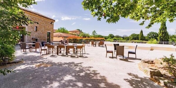 An outdoor patio with tables and chairs, a stone building, and a view of the surrounding landscape. - Casa Jouarres - Appartement avec terrasse privée (Azille)