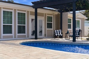 Una piscina con borde de azulejos, un patio cubierto con sillas, y un edificio con ventanas y puerta grandes.
