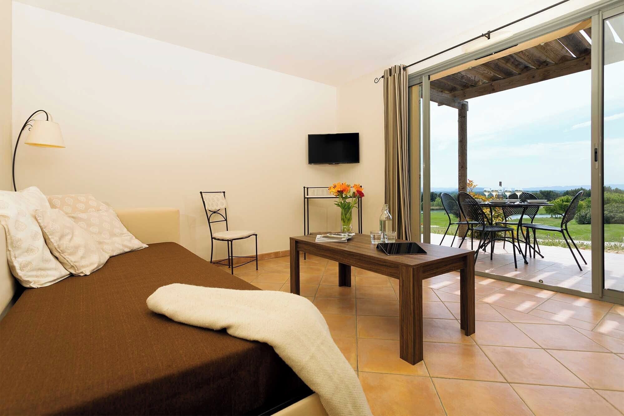 A bedroom with a bed, a TV, a table with a vase of flowers, and a view of a dining area and landscape.