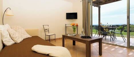A bedroom with a bed, a TV, a table with a vase of flowers, and a view of a dining area and landscape.