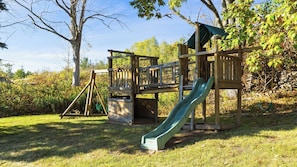 A wooden playground structure with a slide and climbing elements, surrounded by grass and trees. - Mountainview - Guest House by Avantstay Heated Pool + Hot Tub + Games Lake + Cannoe Near Hunter Mountain (Hensonville)
