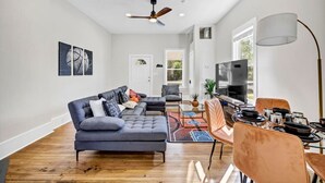 Un salon moderne comprenant un canapé d’angle, une table à manger avec des chaises, un ventilateur de plafond et un téléviseur.