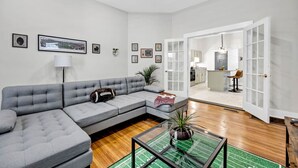 Un salón con un sofá seccional gris, una mesa de centro y un tapete con motivos de fútbol, y una cocina visible a través de una puerta abierta.