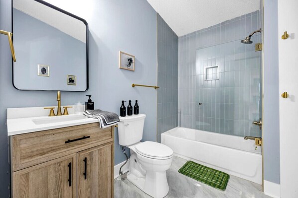 A bathroom with a white toilet, a white sink with gold fixtures, a wooden vanity, a glass shower enclosure, and a green floor mat. - The End Zone | Hot Tub (Columbia)