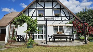 Ein traditionelles Fachwerkhäuschen mit überdachter Terrasse, Gartenmöbeln im Freien und einem Garten.