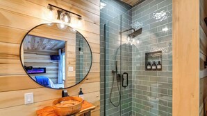 A bathroom with a wooden vanity, a round mirror, a glass shower enclosure, and a small shelf with bottles. - Waterfront cabin-Couples getaway *Private hot tub* (Cherokee)