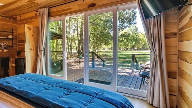 A wooden cabin room with a bed, a sliding glass door to a deck, and a TV mounted on the wall. - Waterfront cabin-Couples getaway *Private hot tub* (Cherokee)