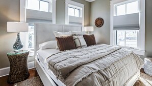 Ein Schlafzimmer mit einem Bett, zwei Nachttischlampen, einer runden Wanduhr und drei Fenstern mit Jalousien.