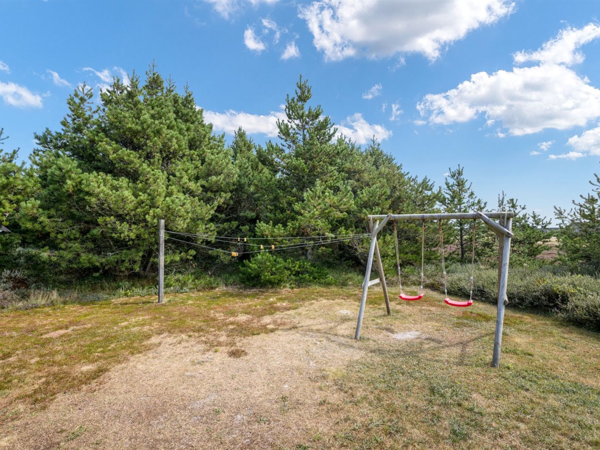 Un terrain de jeux avec des balançoires classiques et une balançoire en corde, entouré d’arbres et d’herbe.