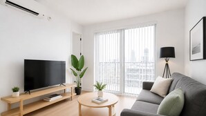 A modern living room with a flat-screen TV, a grey sofa, a round coffee table, and a large window with blinds.