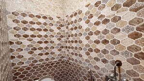 Une salle de bain avec un mur décoré d’un motif imitant la pierre, des toilettes, une douche et une fenêtre avec une grille d’aération.