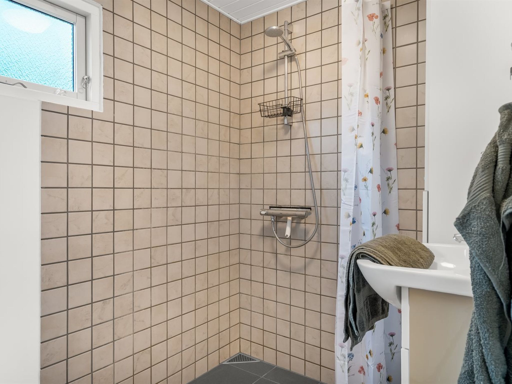 A corner shower with beige tiles, a window, and a floral shower curtain.