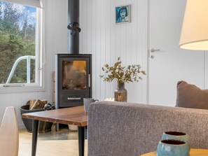 A cozy room with a wood-burning stove, a dining table, a vase with flowers, and a window with a view of trees.