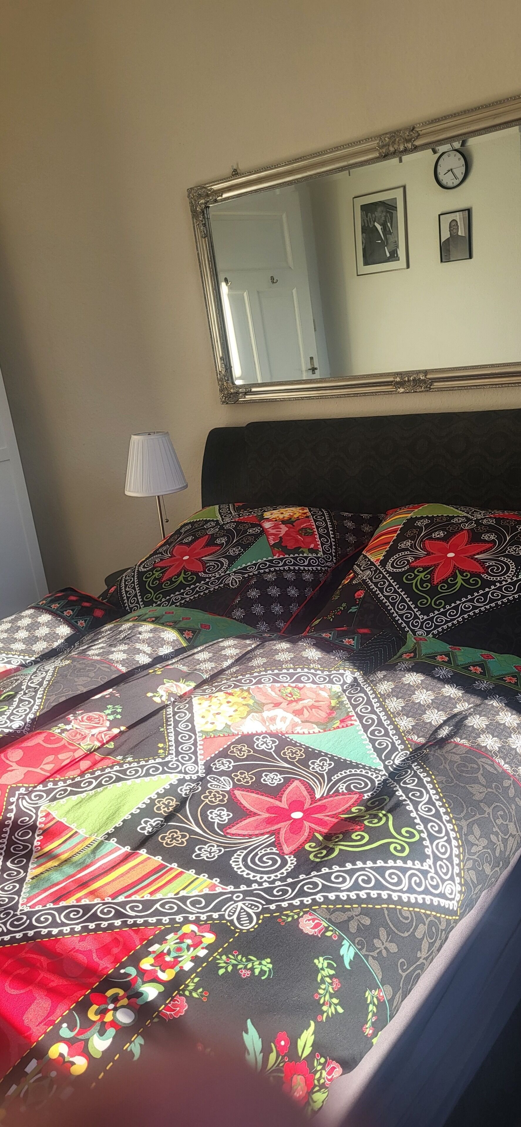 Een bed met een bedsprei met patroon, een bedlamp, een spiegel en een klok aan de muur.