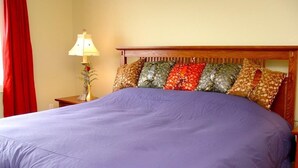 A bed with a purple bedspread, a wooden headboard, and a nightstand with a lamp and a vase. - Island Sunset Resort (Belle Côte)