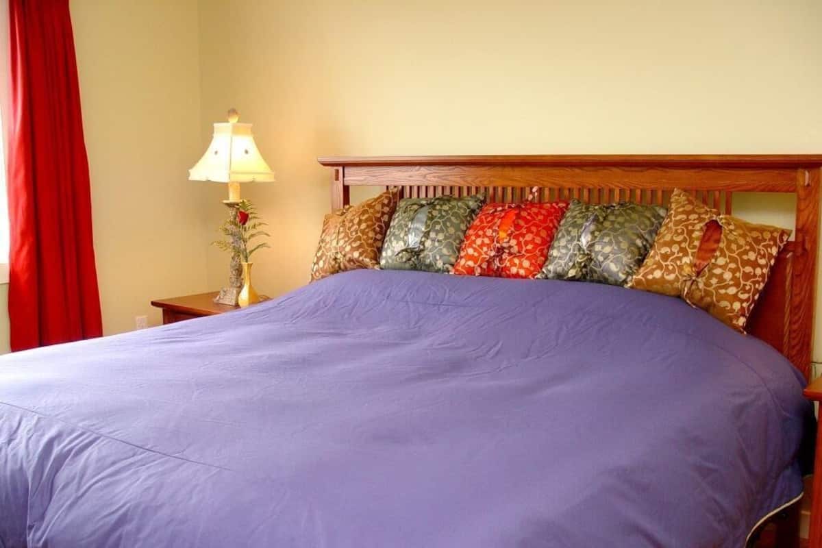 Una cama con cubrecama morado, cabecera de madera y mesita de noche con lámpara y un florero.