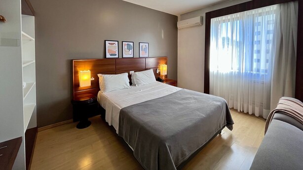 A hotel room with a bed, bedside tables, a headboard, a window with curtains, and a wall-mounted air conditioner. - Apto 606 (Joinville)