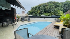A swimming pool with lounge chairs and a table, surrounded by a wooden deck and a brick wall. - Apto 606 (Joinville)