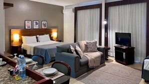 A hotel room with a bed, a sofa, a TV, a dining area with a table and chairs, and a minibar. - Apto 606 (Joinville)