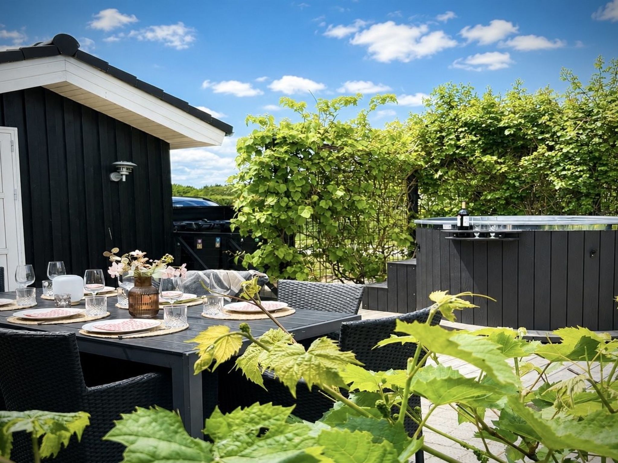 An outdoor dining area with a table set for four, surrounded by greenery and a black fence.