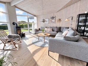 Ein modernes Wohnzimmer mit grauem Sofa, Holzboden, Kamin und einem großen Fenster mit Blick ins Freie.