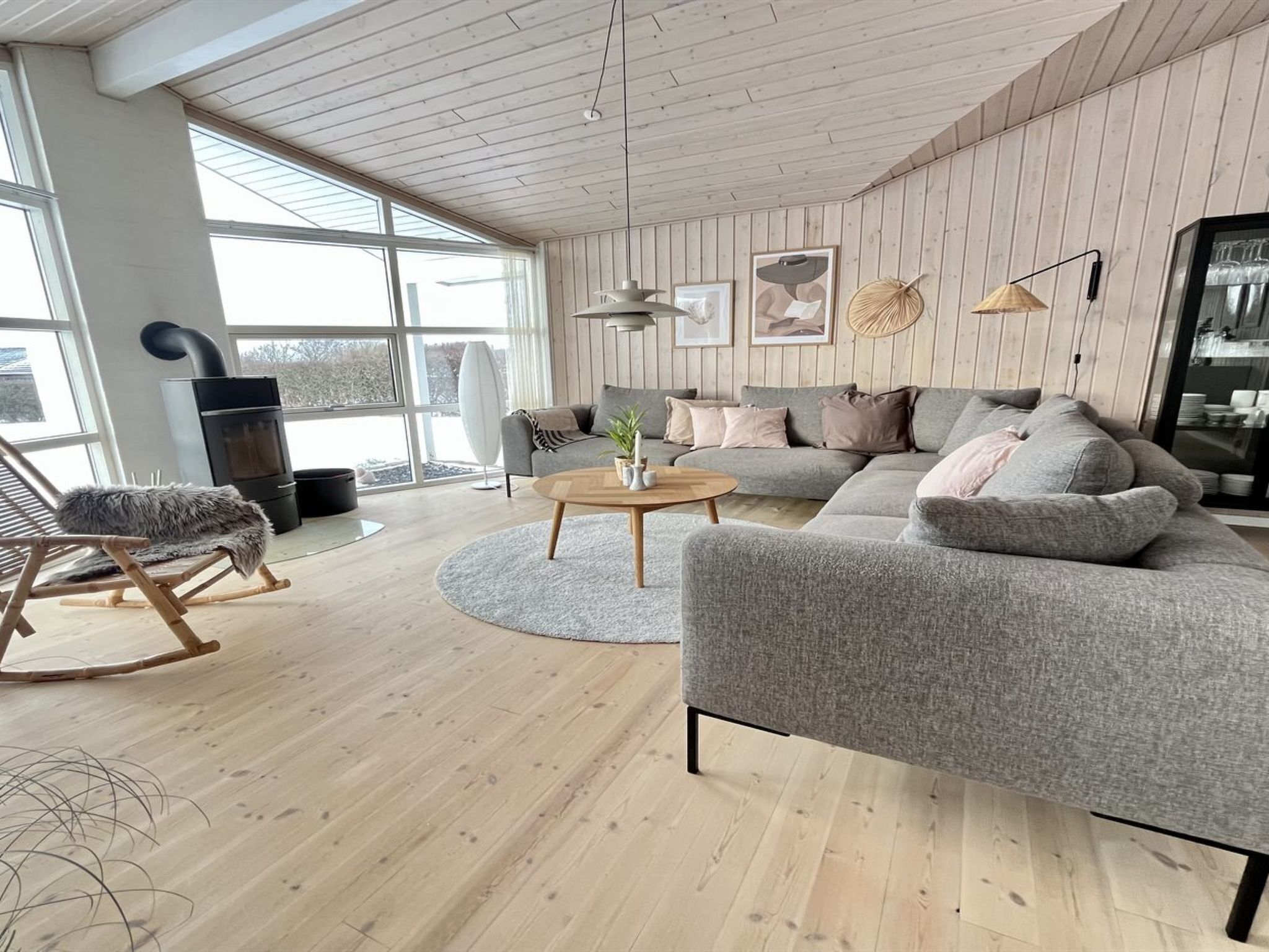 Una sala moderna con un sofá gris, un sillón mecedor de madera, una mesa de centro redonda y un ventanal grande.