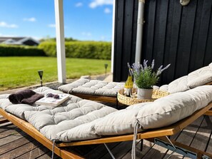 Eine Holzterrasse mit zwei gepolsterten Liegestühlen, einem kleinen Tisch mit einem Blumentopf und einer Weinflasche.