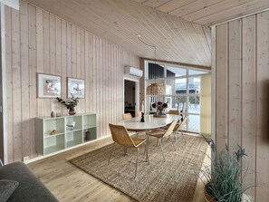 Ein modernes Interieur mit Holzwänden, einem Essbereich mit Stühlen und Tisch, einem weißen Schrank mit Regalen und einem großen Fenster.