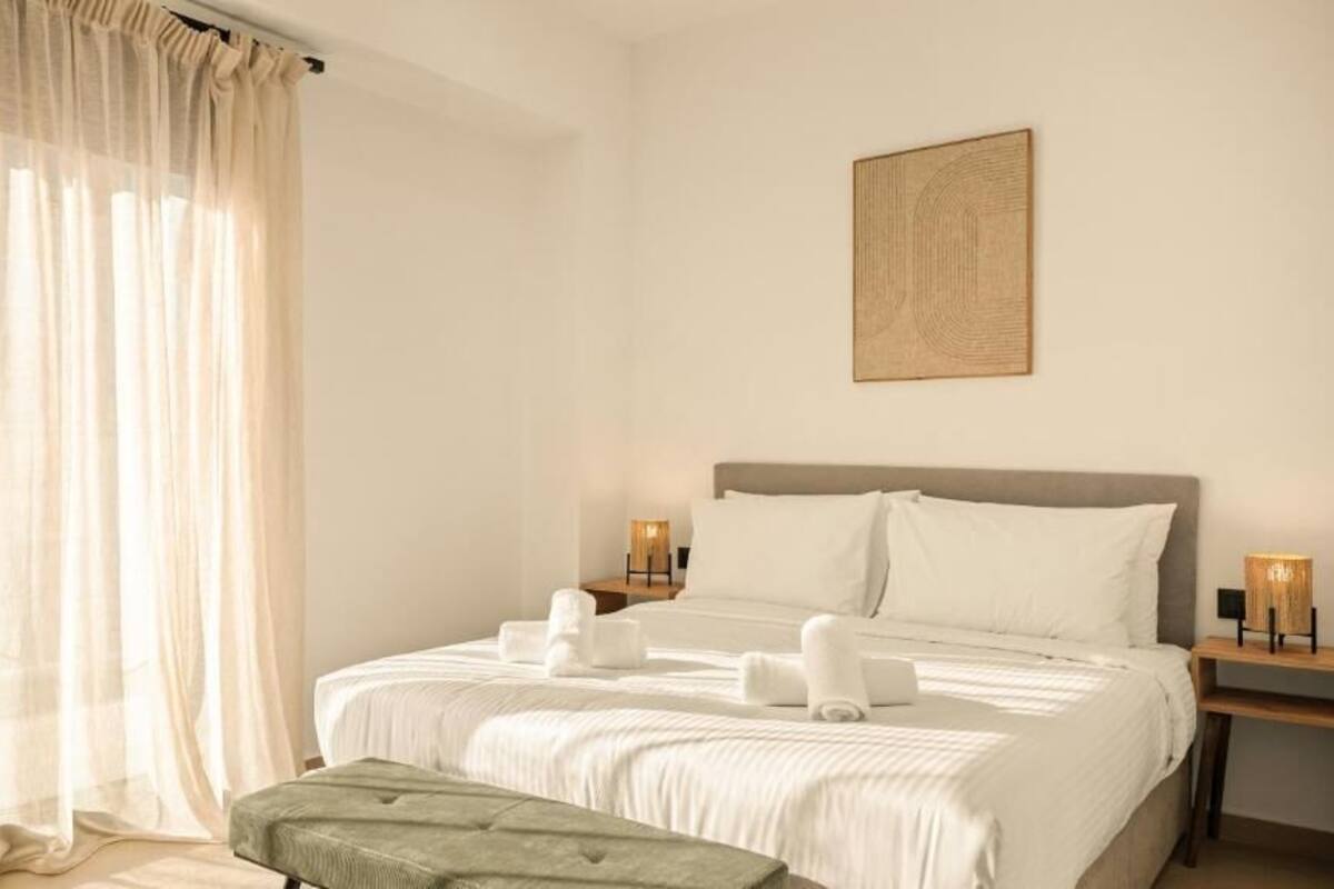 Une chambre d’hôtel avec un lit, une table de chevet avec une lampe, un banc et des rideaux légers.