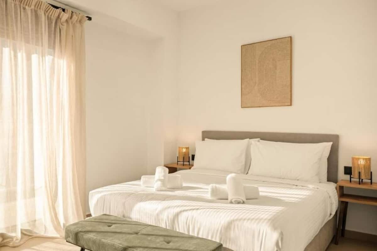A hotel room with a bed, a bedside table with a lamp, a bench, and sheer curtains.