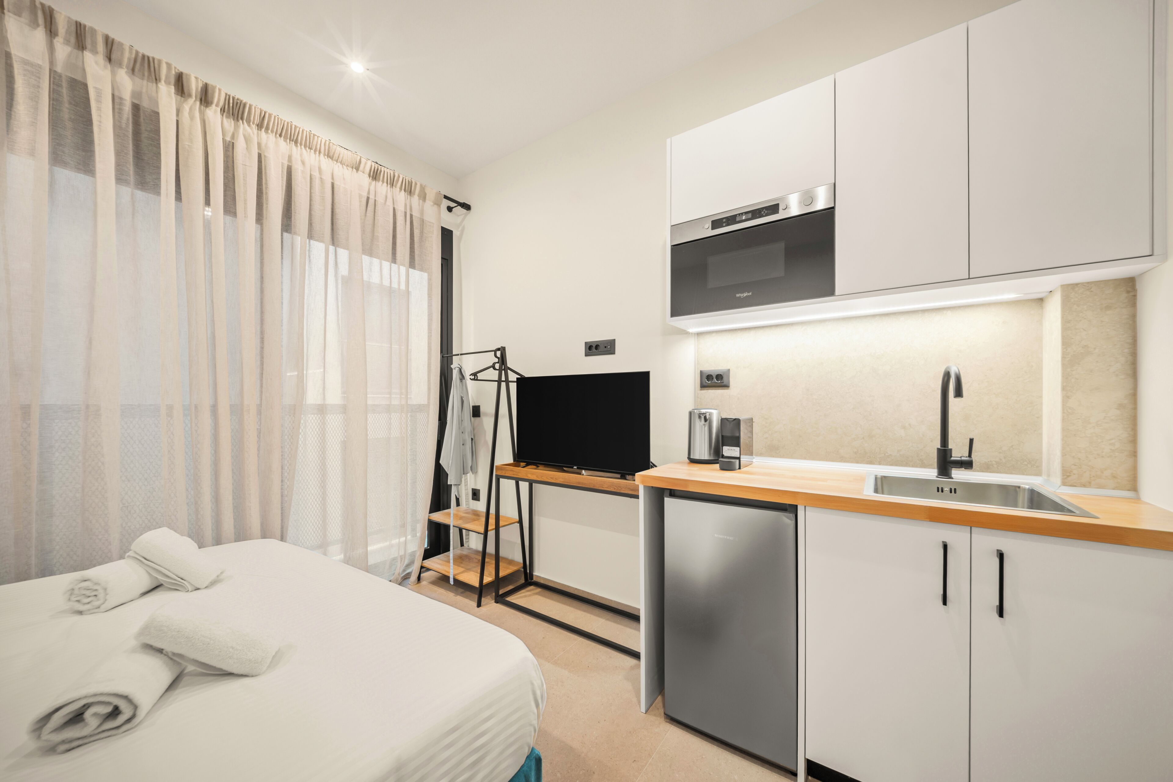 A modern hotel room with a bed, a television, a small kitchenette, and a window with sheer curtains.