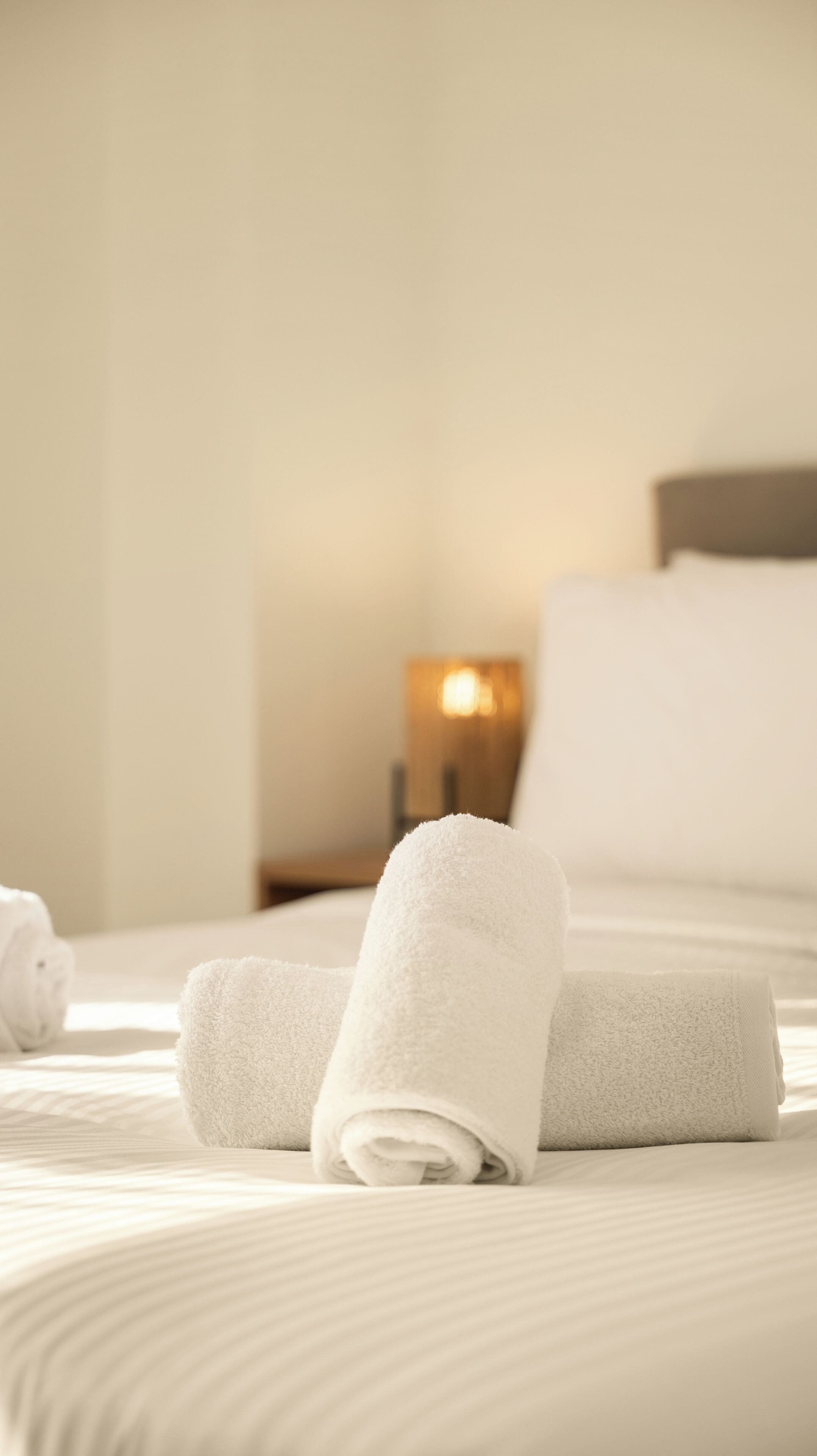 A neatly made bed with white linens and rolled towels, a bedside table with a lamp, and a wooden headboard.