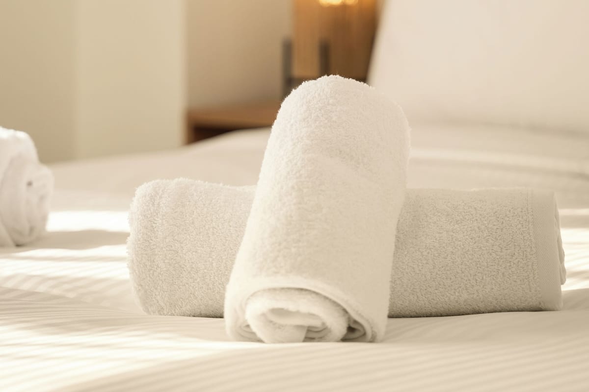 A neatly made bed with white linens and rolled towels, a bedside table with a lamp, and a wooden headboard.