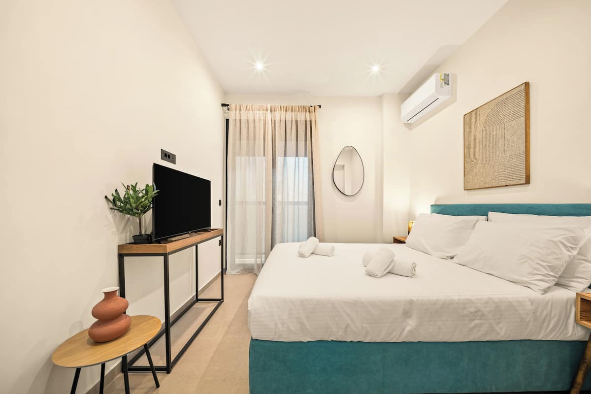 A hotel room with a bed, a TV, a plant, and a painting on the wall.