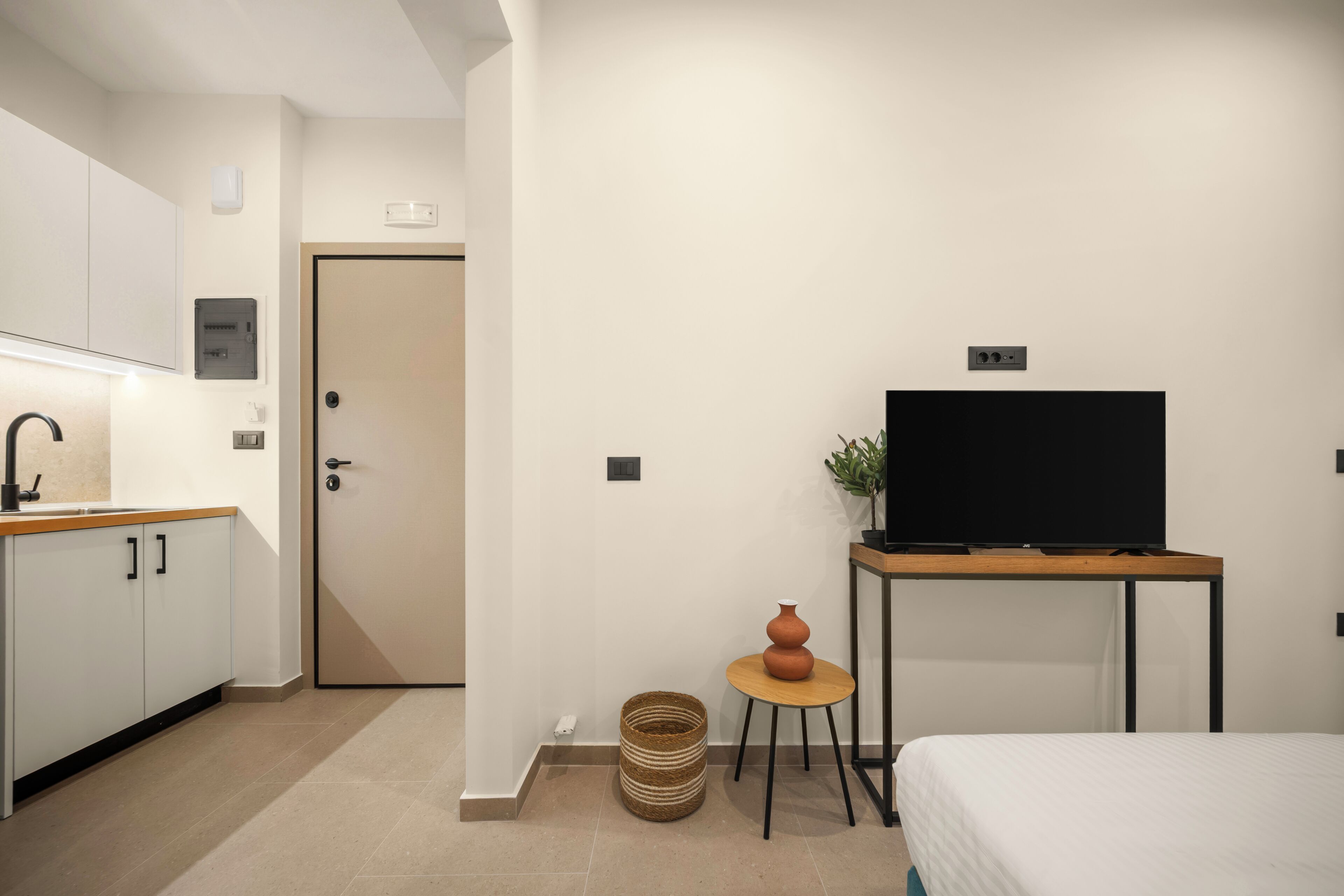 Moderni hotellihuone, jossa on keittotila, televisio puupöydällä ja ovi, joka johtaa toiseen huoneeseen.