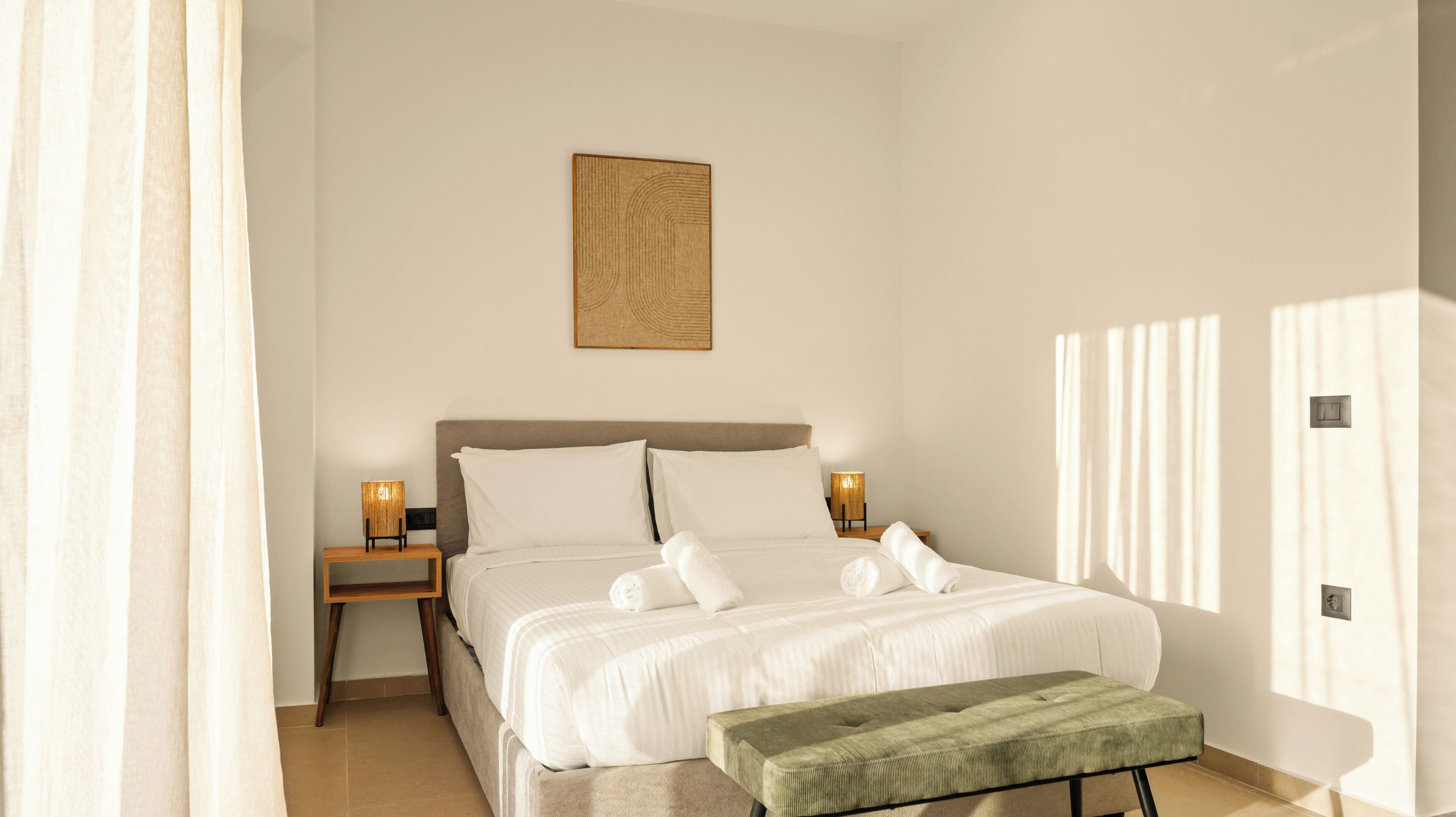 A bedroom with a bed, bedside lamps, a green bench, and a wall-mounted artwork.
