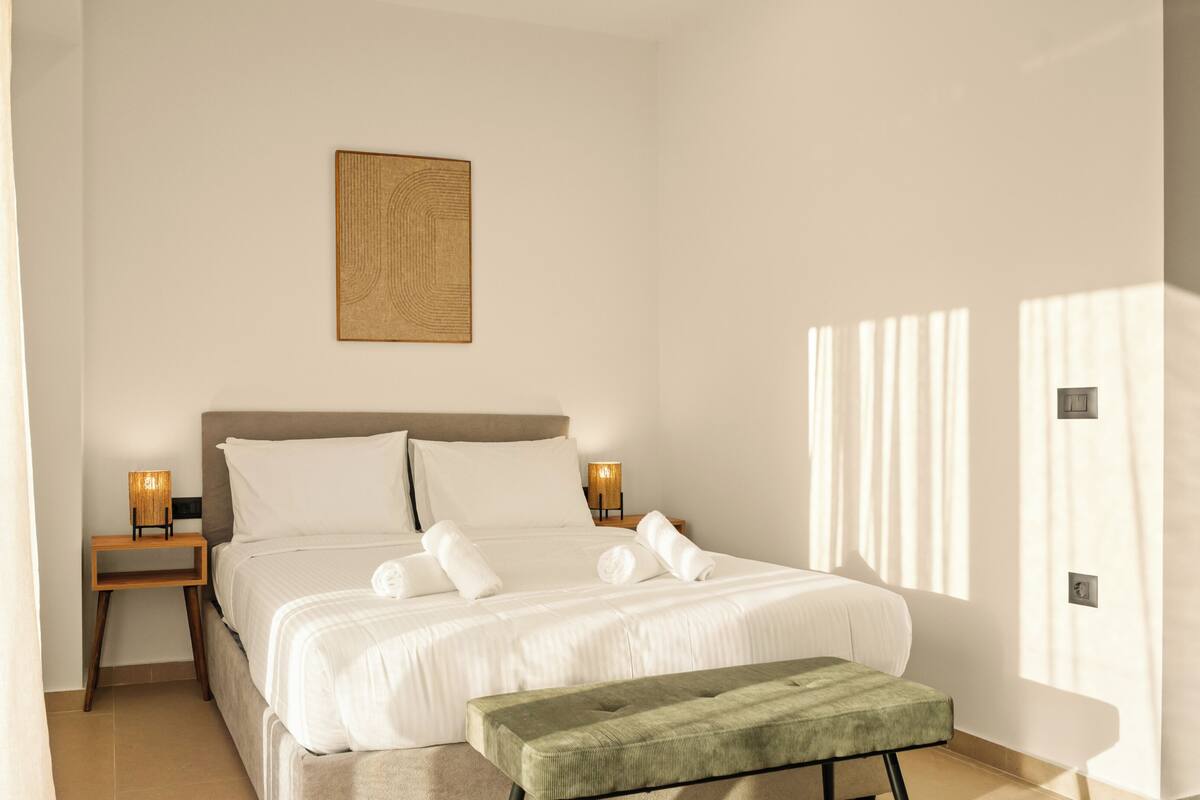 Une chambre à coucher avec un lit, des lampes de chevet, un banc vert et une œuvre d’art accrochée au mur.