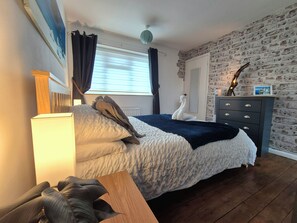 A bedroom with a bed, a dresser, a lamp, a wall-mounted radiator, a window with curtains, and a brick-patterned wall. - Home from Home - Entire 1 bed House, Parking, Sky TV, Fast Fibre, London Train. (Snodland)
